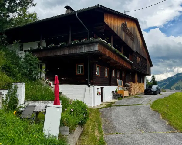 Drewniana austeria w górach, z balkonami ozdobionymi kwiatami. Obok stoi czarny samochód, a w oddali widać zalesione wzgórza.