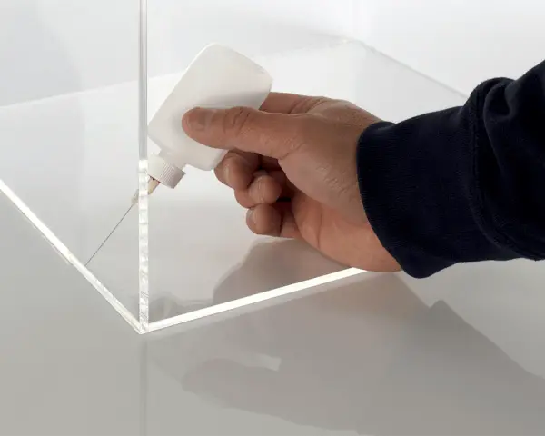 A hand applies adhesive to the corner of a clear plastic box, demonstrating precise gluing of plastic for a clean finish.