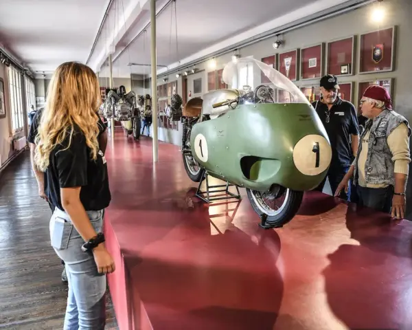 Un raduno moto Guzzi in un museo. Una moto da corsa verde con il numero 1 in primo piano, ammirata da visitatori.