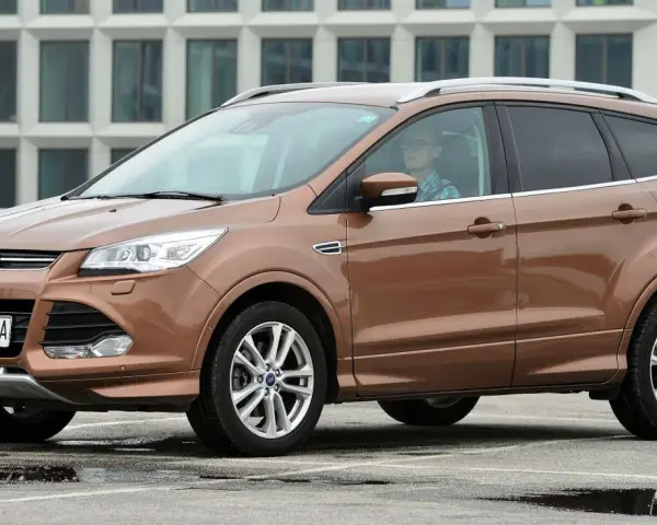 Brązowy Ford Kuga 2019 z kierowcą w okularach, zaparkowany na mokrym parkingu przed nowoczesnym budynkiem.