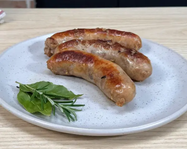 Tre salsicce dorate e succulente, appena cotte in padella, riposano su un piatto con rametti di rosmarino e alloro.