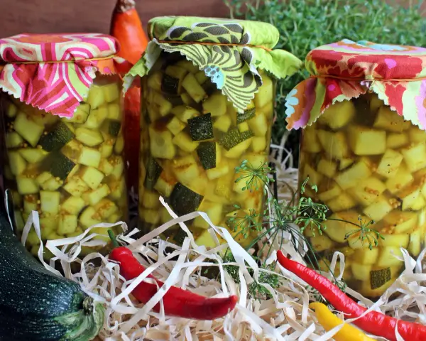Słoiki z cukinią w zalewie słodko-kwaśnej z curry, ozdobione kolorowymi materiałami. Obok leżą świeże cukinie i papryczki chili.