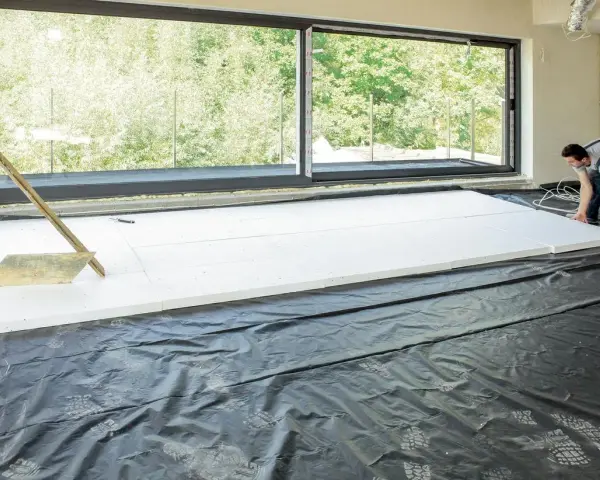 Pracownik układa płyty styropianowe do ocieplenie stropu betonowego. Duże okno wychodzi na zieleń.