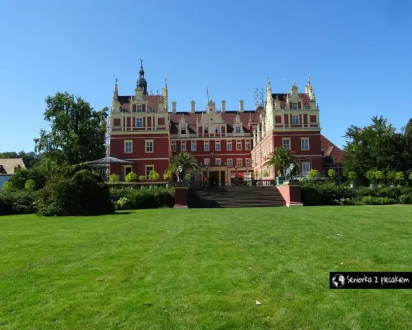 Majestatyczny pałac w parku muzakowskim, otoczony zielonym trawnikiem i drzewami, pod błękitnym niebem.