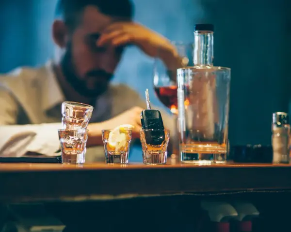 Ein Mann mit Kopfschmerzen sitzt am Tresen, neben Autoschlüsseln in einem Schnapsglas. Alkohol am Steuer unter 21 ist eine Gefahr.