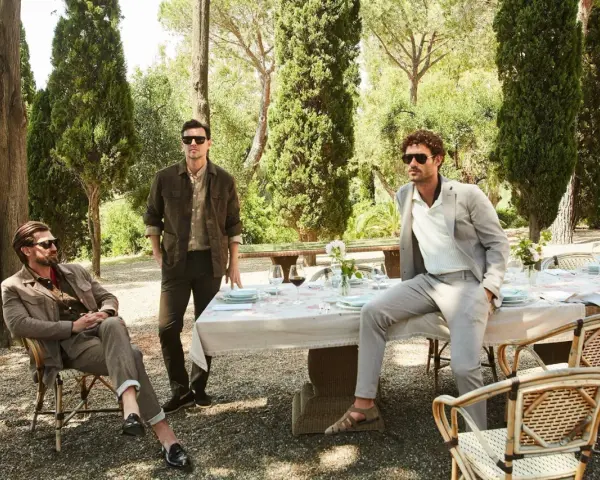 Uomini eleganti in un giardino, con un uomo seduto che indossa un completo marrone e un altro in piedi con un completo grigio. Il pantalone, in entrambi i casi, definisce lo stile.