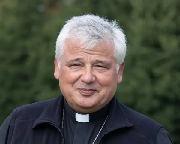 Uśmiechnięty jałmużnik papieski w sutannie i czarnym polarze, na tle zieleni.