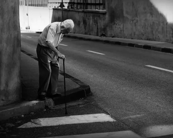 Une photo dans la rue : un vieil homme se penche sur sa canne, le regard perdu, au bord d'une route déserte.