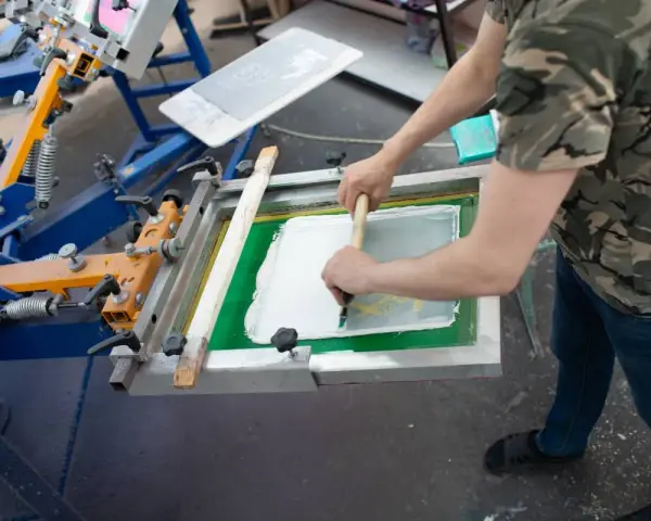 Proceso de serigrafía que es: un operario aplica tinta blanca sobre una pantalla para estampar.