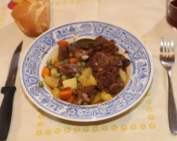 Un plato de corzo guisado con patatas, zanahorias y guisantes, acompañado de pan y vino tinto. ¡Una de las mejores recetas de corzo guisado!
