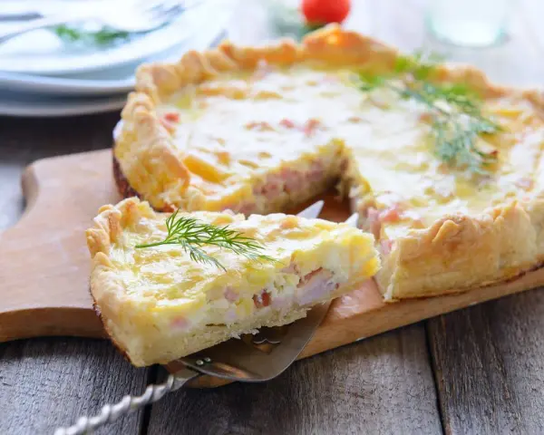 Una fetta di torta salata prosciutto e formaggio, guarnita con aneto, su un tagliere di legno.