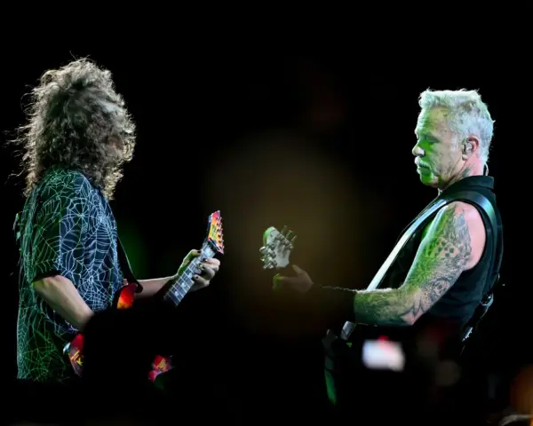 James Hetfield, z charakterystycznym wąsem i wytatuowanymi ramionami, gra na gitarze. Obok niego drugi muzyk z burzą włosów.