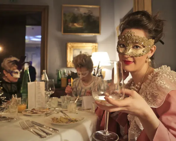 Elegancka kobieta w złotej masce i perłach świętuje karnawał 2026. Wznosi kieliszek wina, otoczona innymi gośćmi w maskach.