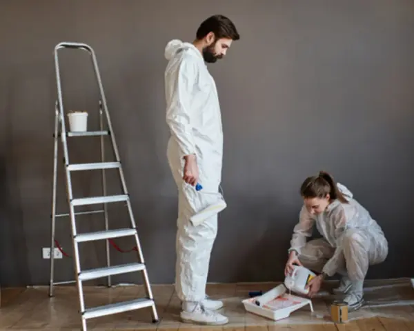 Pareja aprendiendo a pintar su hogar. Él sostiene un rodillo, ella vierte pintura. Hay cajas y una escalera.
