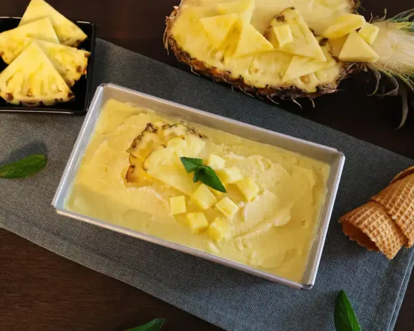 Erfrischendes Ananas Eis in einer Metallform, garniert mit Ananasstücken und Minze. Daneben liegen Ananasspalten und eine Waffel.