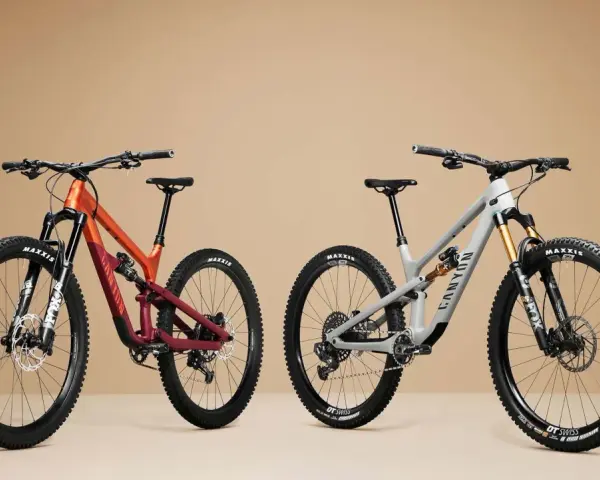 Two full-suspension mountain bikes, one orange and maroon, the other grey, are displayed side-by-side, showcasing the 27.5 vs 29 wheel size debate.
