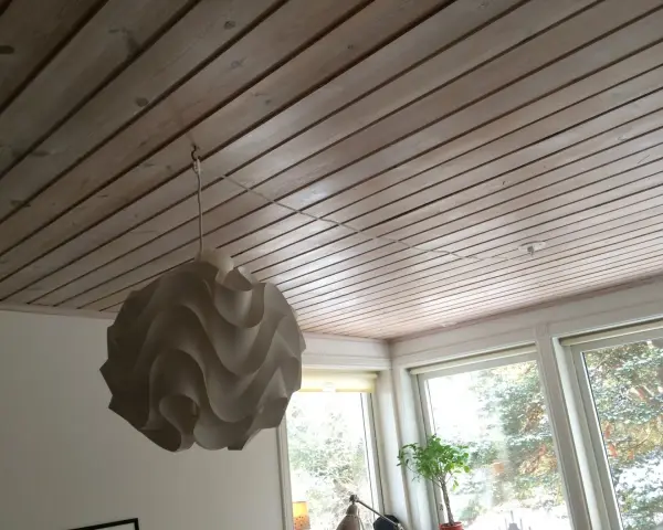 En vit lampskärm hänger från ett tak av träpanel. En tunn sladd dras i taket för att ansluta lampan.