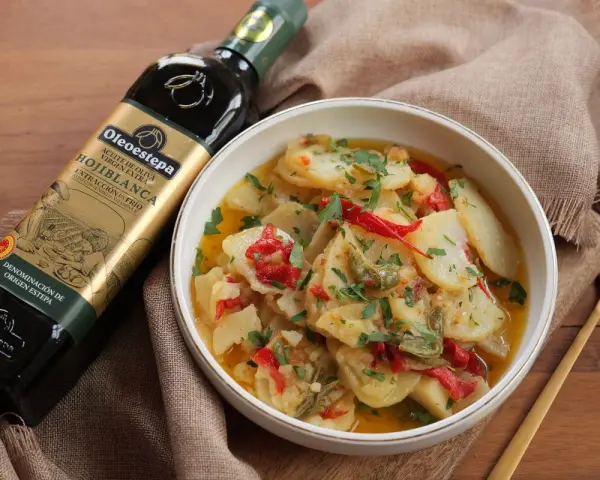 Un tazón de patatas a lo pobre, con pimientos rojos y verdes, perejil picado y una botella de aceite de oliva virgen extra.