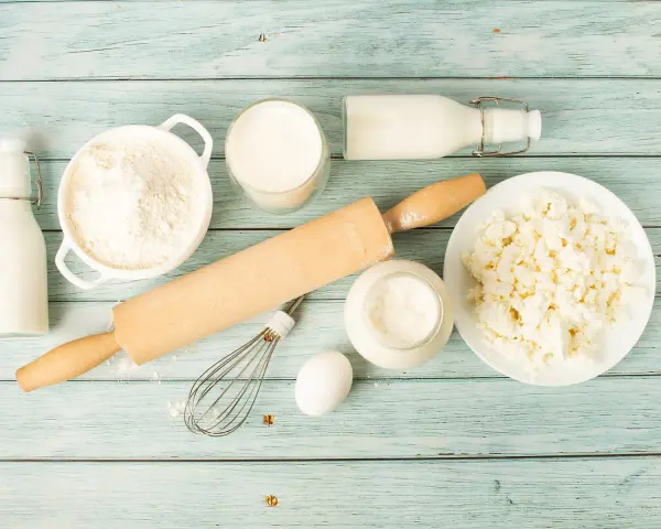 Zutaten für butter ersatz backen: Mehl, Milch, Quark, Ei, Schneebesen und Nudelholz auf blauem Holzuntergrund.