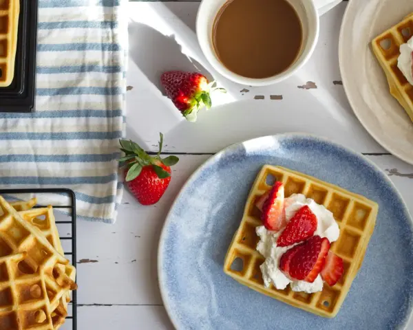 Frische belgische Waffeln mit Erdbeeren und Sahne auf einem blauen Teller, daneben eine Tasse Kaffee und weitere Waffeln auf einem Gitter.