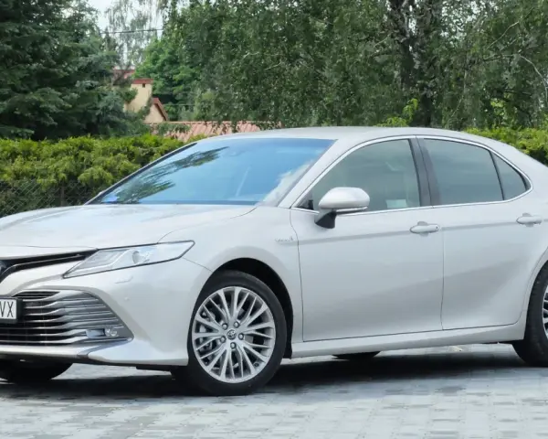 Srebrna Toyota Camry 2.5 Hybrid stoi na wybrukowanym podjeździe, otoczona zielenią.