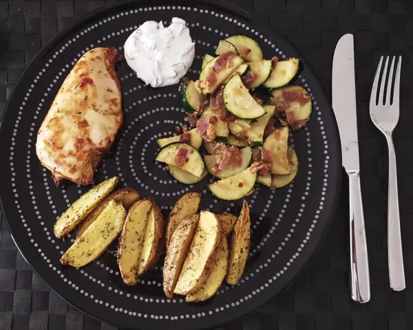 Ein Teller mit Hähnchenbrust, Kartoffelspalten und Zucchinigemüse, perfekt für Airfryer Rezepte. Dazu ein Klecks Dip.