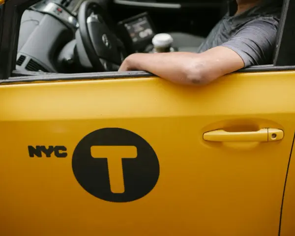 Kierowca żółtej taksówki NYC z logo "T" w kole. Zastanawiasz się, ile zarabia taksówkarz w tym mieście?