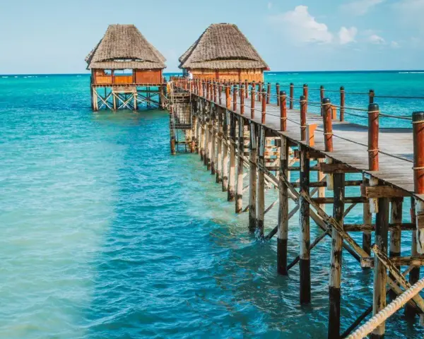 Drewniany pomost prowadzi do chat na palach nad turkusową wodą. Zanzibar, pora deszczowa, ale słońce świeci.