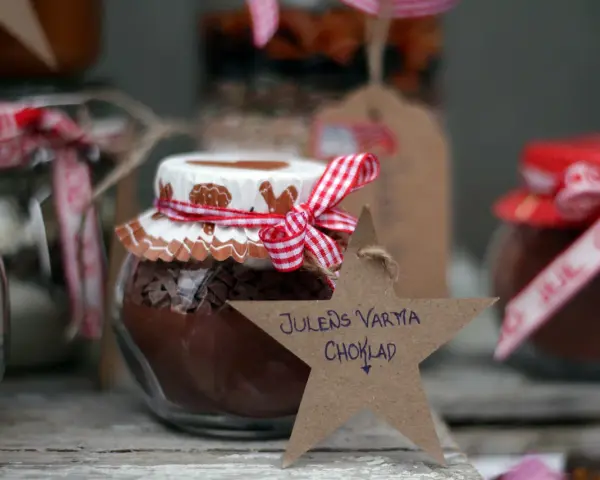Burkar med ingredienser för varm choklad jul, dekorerade med röda band och pepparkaksfigurer. En stjärna med texten "Julens varma choklad" hänger framför.