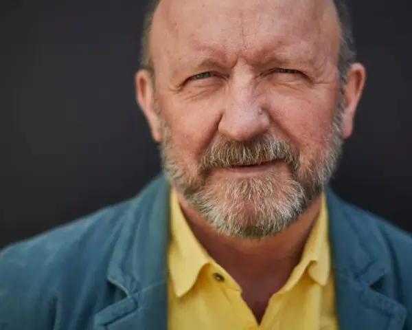 Artur Barciś w żółtej koszuli i niebieskiej marynarce, z siwą brodą, patrzy w obiektyw.