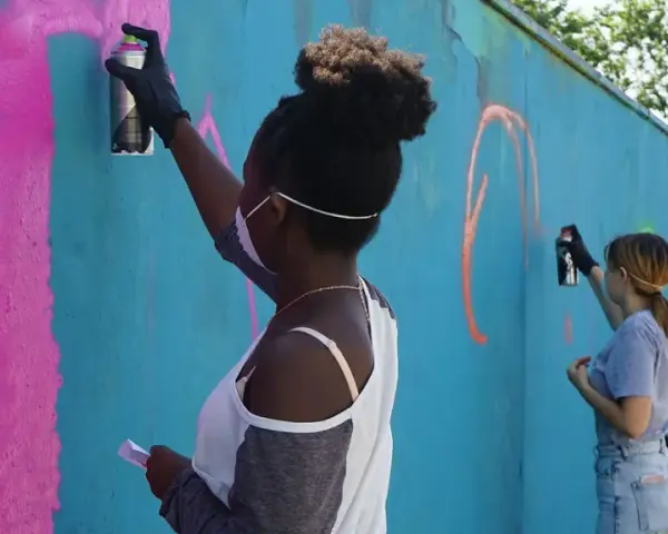 Ungdomar målar graffiti på en blå vägg. En flicka med afrohår sprayar rosa färg.