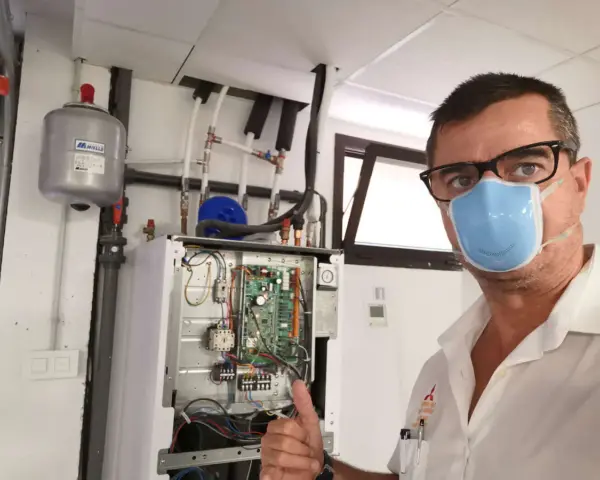 Técnico con mascarilla realiza mantenimiento aerotermia. Equipo abierto, circuito visible. Pulgar arriba.