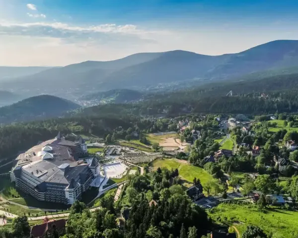 Malownicze Karkonosze latem: rozległy kompleks hotelowy otoczony zielenią, z widokiem na góry i miasteczko. Idealne miejsce na wypoczynek i odkrywanie atrakcji.