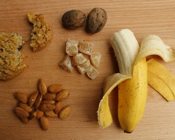 Na drewnianym blacie leżą orzechy, migdały, kawałek ciastka, kandyzowany imbir i banan. To świetne węglowodany złożone produkty.