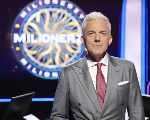 Hubert Urbański w garniturze, na tle koła z napisem "Milionerzy". Co dajemy komuś na drogę milionerzy? Wiedzę i odwagę!