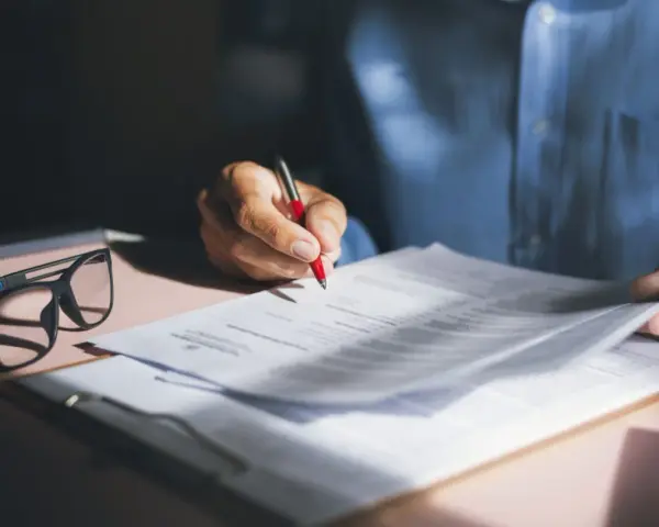 Persona revisa y firma una nota de encargo inmobiliaria, con gafas y bolígrafo rojo sobre documentos.