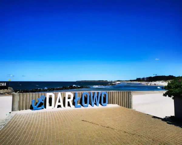 Nadmorskie miasto nad Wieprzą, Darłowo, wita turystów na piaszczystej plaży. Błękitne niebo i morze tworzą idealne tło dla napisu.