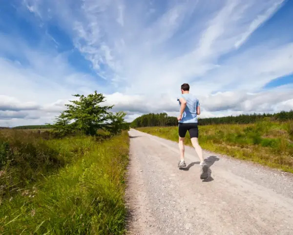 Uomo corre su strada sterrata, ideale per la preparazione 10 km principianti. Cielo azzurro con nuvole bianche.