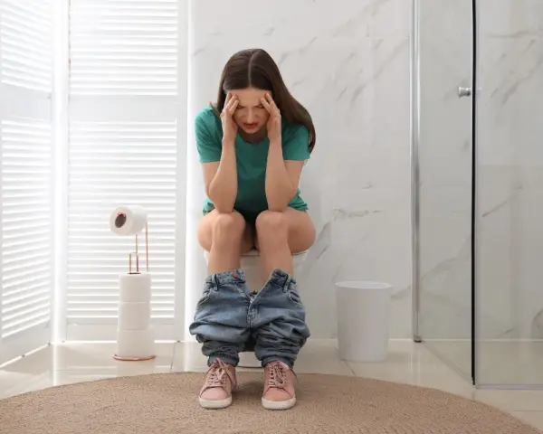 Donna seduta sul water con le mani sulla testa, visibilmente sofferente. Sembra che abbia mangiato troppo e ora si senta male, come se avesse "feci a palline".