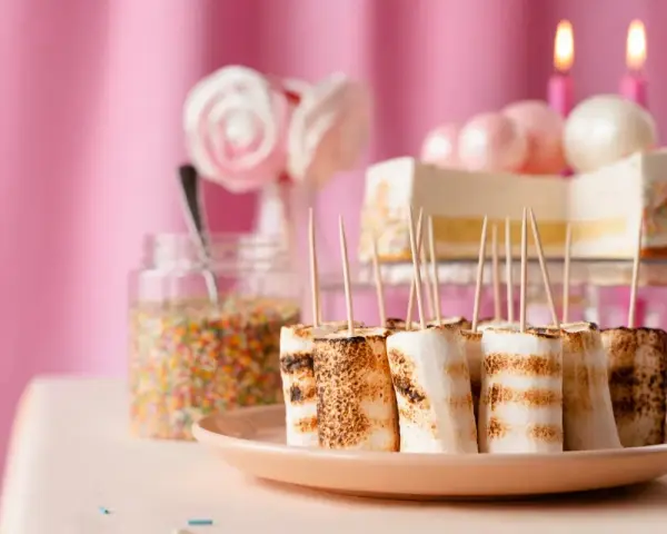Rostade marshmallows på spett, perfekt för roliga recept att baka till kalaset. En burk strössel och en tårta i bakgrunden.