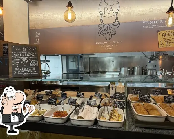 A display case filled with small dishes of food, with a chalkboard menu and a restaurant logo in the background.
