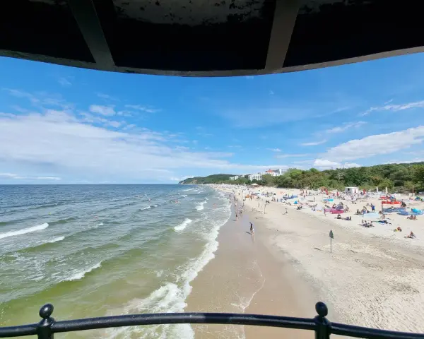 Słoneczna plaża z tłumem turystów, gdzie jest najcieplejsze morze. Woda faluje, a na piasku rozłożone są parawany i ręczniki.