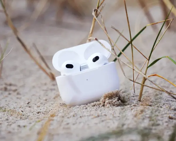 Weiße AirPods im geöffneten Ladecase liegen im Sand, umgeben von Gräsern.