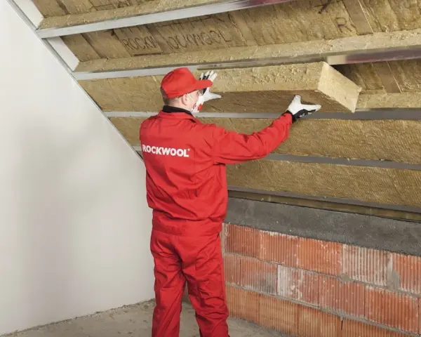 Pracownik w czerwonym kombinezonie Rockwool montuje izolację na ścianie szczytowej poddasza.