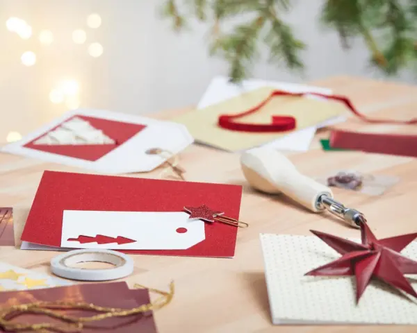 Förberedelser för att måla julkort. Rött glitterpapper, vita etiketter med julgransmotiv och en stjärnformad gemklämma ligger på ett träbord.