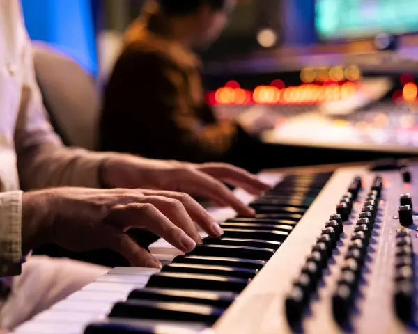 Hände spielen Synthesizer-Tasten, während im Hintergrund ein Tontechniker an einem Mischpult arbeitet. Die Szene fängt die Entstehung eines neuen ep album ein.