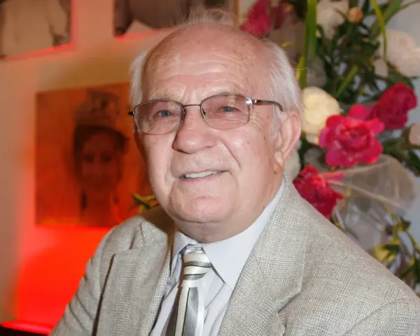 Uśmiechnięty Jerzy Antczak, reżyser znany z niezapomnianych filmów, w eleganckim garniturze, na tle bukietu kwiatów i portretu.