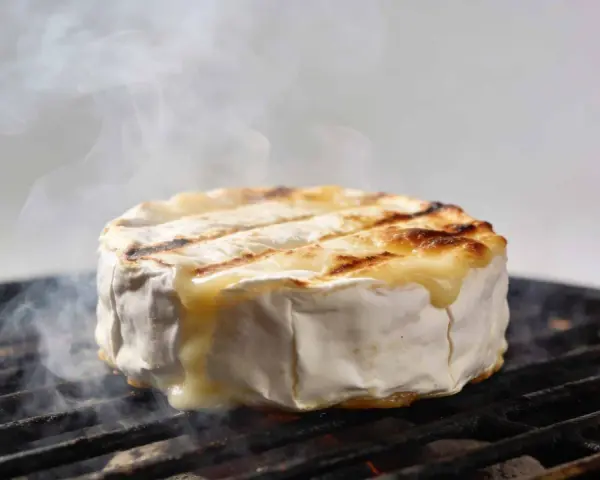 Un camembert au barbecue, fondant et fumant, cuit sur une grille. La croûte blanche se craquelle, révélant un cœur doré et coulant.