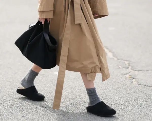 Persona camina con zuecos negros y calcetines grises, llevando un bolso negro y un abrigo beige.