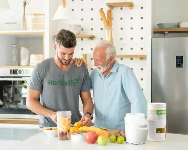 Två män i ett kök förbereder en smoothie med frukt. En burk med vegetariskt protein står på bänken.